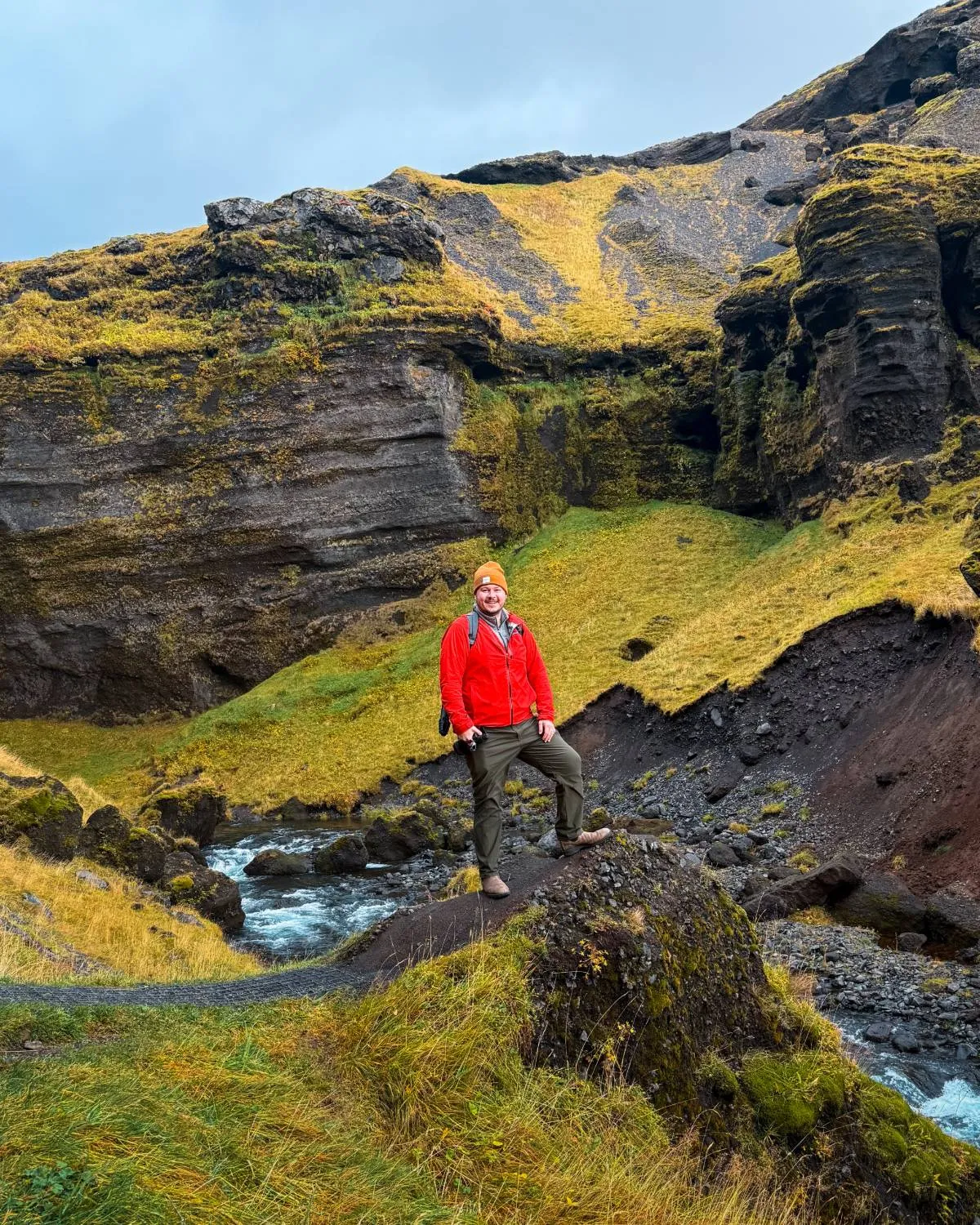 Andy Feliciotti in a red jacket and orange beanie, standing on a rock above a river in a mossy canyon in Iceland